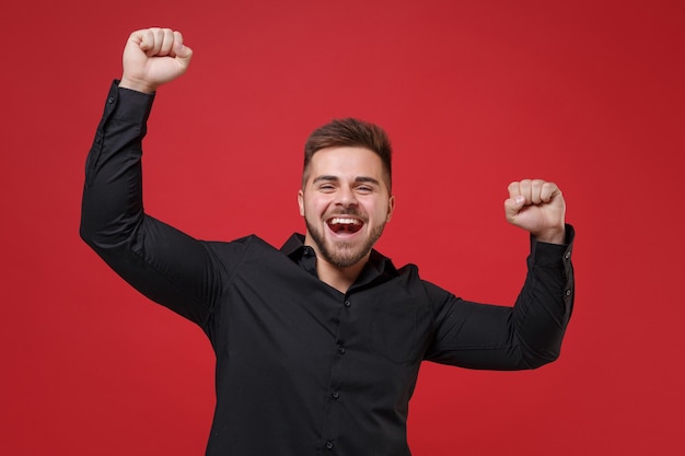 Joyeux jeune homme barbu des années 20 en chemise noire classique posant isolé sur un portrait en studio de fond rouge vif. Concept de style de vie d'émotion sincère de personnes. Maquette de l'espace de copie. Serrer les poings comme vainqueur.