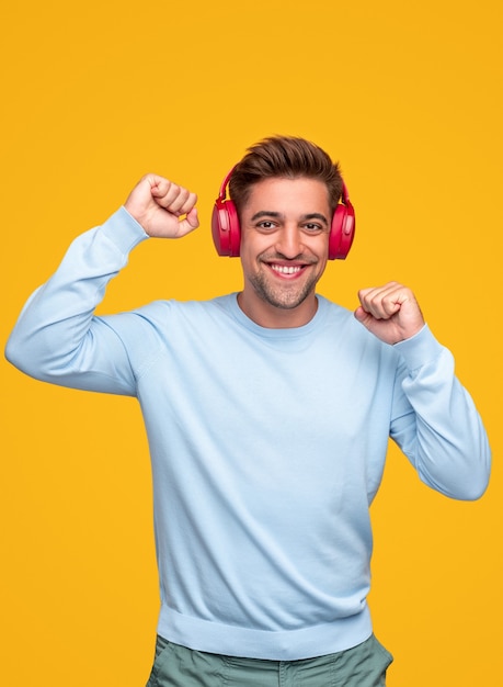 Joyeux jeune homme barbu en tenue décontractée s'amusant et dansant tout en écoutant de la musique avec des écouteurs sans fil sur fond jaune