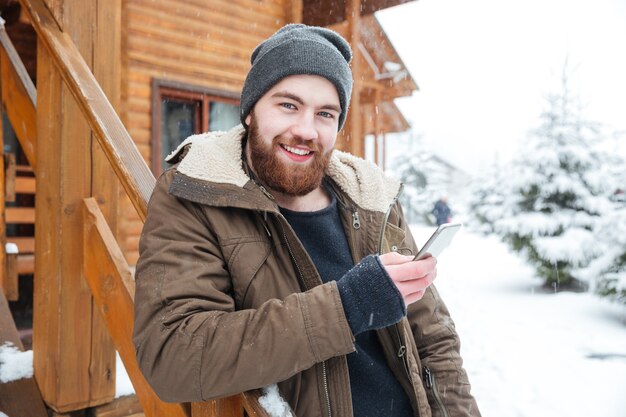 Joyeux jeune homme barbu utilisant un smartphone à l'extérieur en hiver