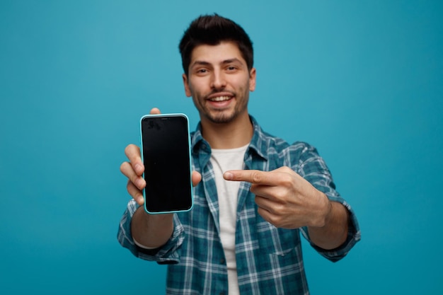 Joyeux jeune homme étirant le téléphone portable vers la caméra en le pointant en regardant la caméra isolée sur fond bleu