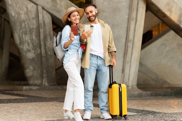Joyeux jeune homme européen avec une dame à barbe détenant un passeport prenant un selfie sur un smartphone et profitant d'un voyage
