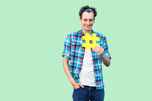 Joyeux jeune homme optimiste et joyeux en chemise à carreaux tenant un grand signe de hashtag jaune, concept de médias sociaux. Intérieur, isolé, tourné en studio, espace copie, fond vert