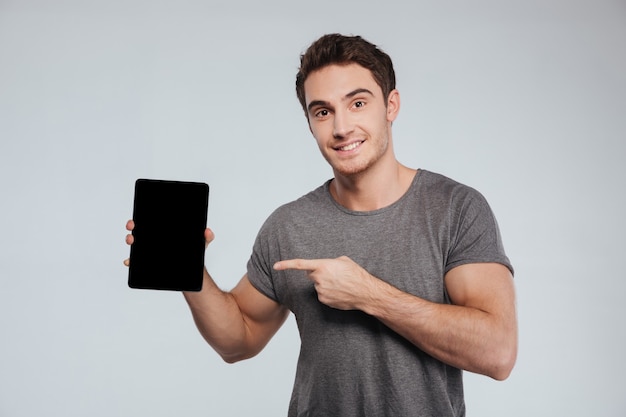 Joyeux jeune homme pointant le doigt sur une tablette à écran blanc sur fond gris