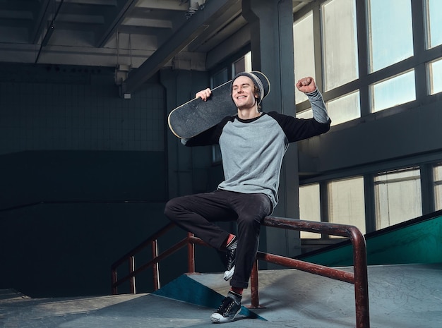 Joyeux jeune homme portant une chemise et un chapeau tenant sa planche, assis sur un rail de broyage dans le skatepark à l'intérieur.