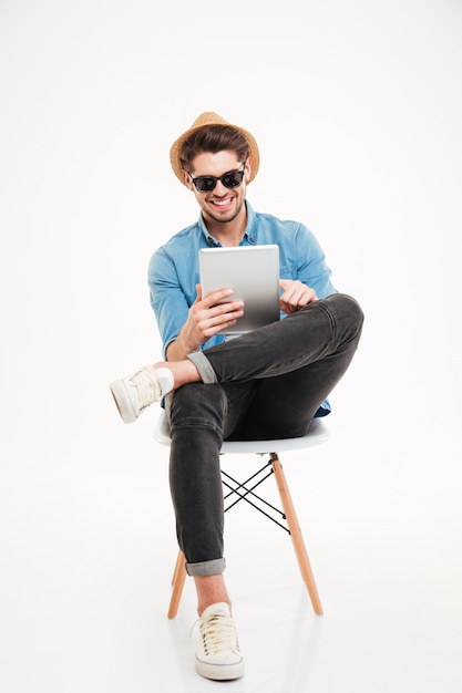 Joyeux jeune homme séduisant portant des lunettes assis et tenant une tablette isolée