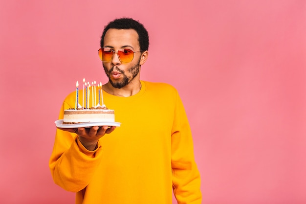 Joyeux jeune homme soufflant des bougies sur un gâteau d'anniversaire isolé sur rose.