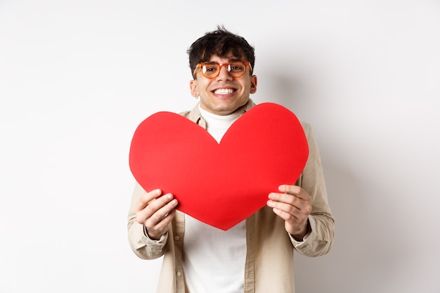 Joyeux jeune homme souriant et l'air ravi devant la caméra, montrant une grande découpe de coeur rouge le jour de la Saint-Valentin, faisant un cadeau romantique à l'amant, fond blanc.