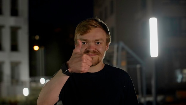 Joyeux jeune homme souriant avec son pouce en l'air dans la rue la nuit médias homme caucasien posant dans