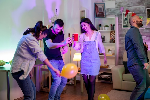 Joyeux jeune homme tenant une bouteille de bière en dansant avec ses amis lors d'une fête avec de la musique disco et des ballons.