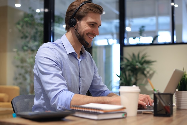 Joyeux jeune opérateur de téléphone de soutien dans le casque, sur le lieu de travail tout en utilisant un ordinateur portable, un service d'aide et un concept de centre d'appels de consultation client.