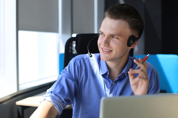 Joyeux jeune opérateur de téléphone de soutien dans le casque, sur le lieu de travail tout en utilisant un ordinateur portable, un service d'aide et un concept de centre d'appels de consultation client.