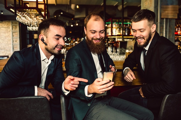 De joyeux jeunes hommes d'affaires positifs et gentils sont assis dans le bar. Ils regardent le téléphone. Le premier gars le pointe. Ils sourient.