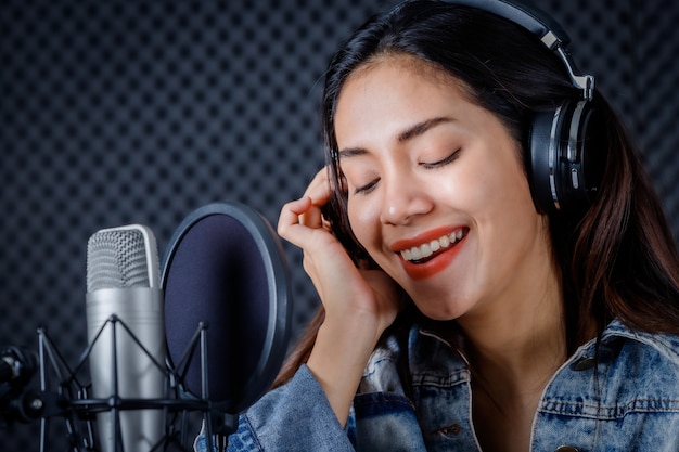 Joyeux joyeux assez souriant du portrait d'une jeune chanteuse asiatique portant des écouteurs enregistrant une chanson devant un microphone dans un studio professionnel