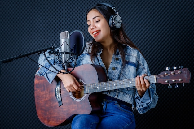 Joyeux joyeux assez souriant du portrait d'une jeune chanteuse asiatique portant des écouteurs avec une guitare enregistrant une chanson devant un microphone dans un studio professionnel