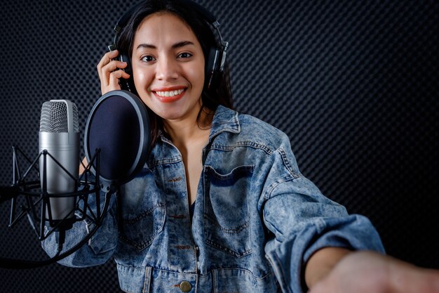 Joyeux joyeux assez souriant du portrait d'une jeune chanteuse asiatique portant des écouteurs prenant un selfie avec un smartphone tout en enregistrant une chanson devant le microphone dans un studio professionnel