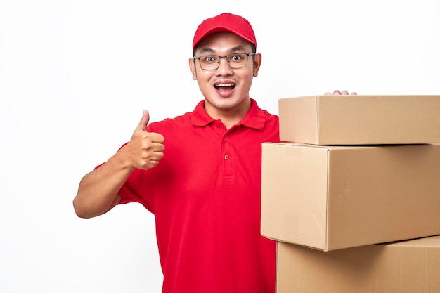 Joyeux livreur souriant en chemise rouge et casquette debout près de tas de boîtes de colis montrent le pouce en signe d'approbation