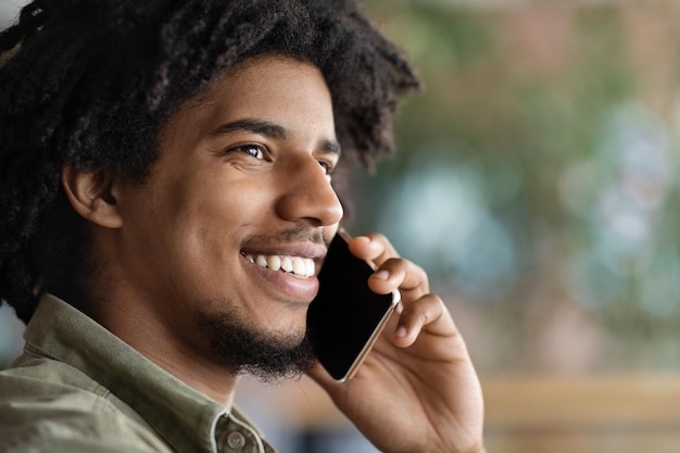 Photo joyeux mec bouclé afro-américain du millénaire parlant par smartphone à l'intérieur du café en gros plan