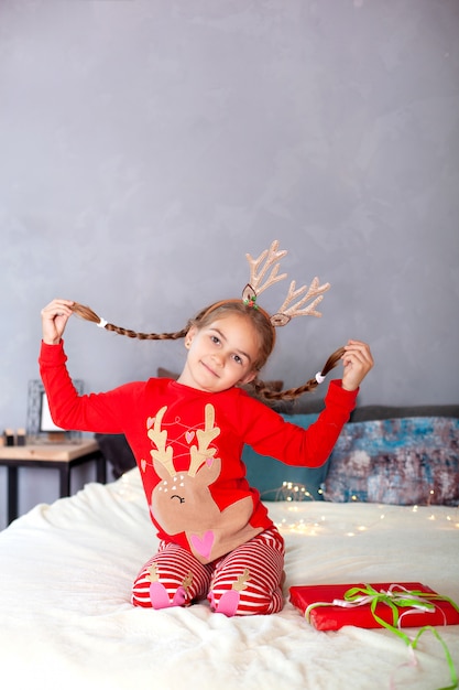 Joyeux Noël. Nouvel An. Petite fille avec des nattes sur la tête avec boîte-cadeau de Noël à la maison. enfant en pyjama rouge et avec des cornes de cerf sur la tête la veille de Noël s'amuse et ouvre le cadeau. Hiver