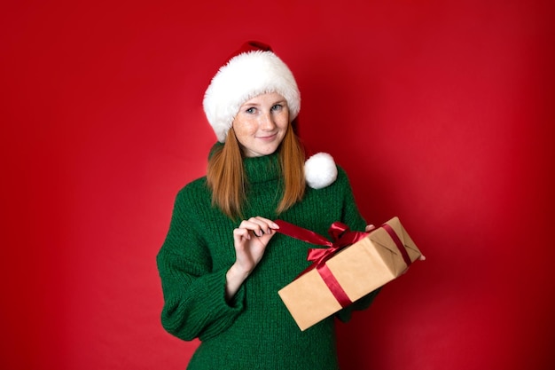 Joyeux Noël Portrait d'une belle jeune adolescente dans un chandail vert tricoté confortable et un chapeau du Père Noël tenant des coffrets cadeaux Le fond rouge est l'endroit pour le texte