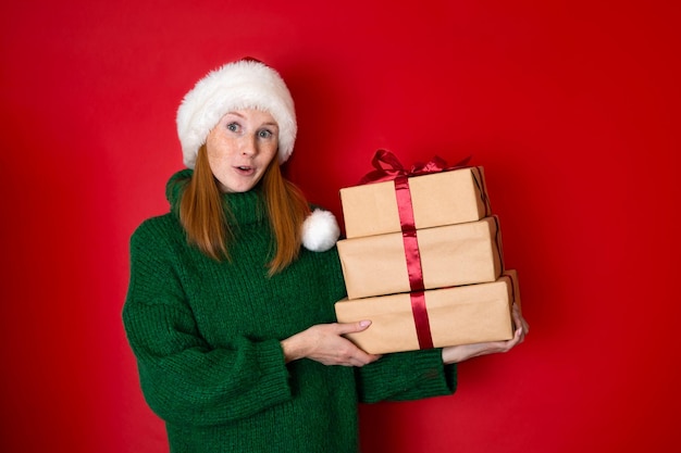 Joyeux Noël Portrait d'une belle jeune adolescente dans un chandail vert tricoté confortable et un chapeau du Père Noël tenant des coffrets cadeaux Le fond rouge est l'endroit pour le texte