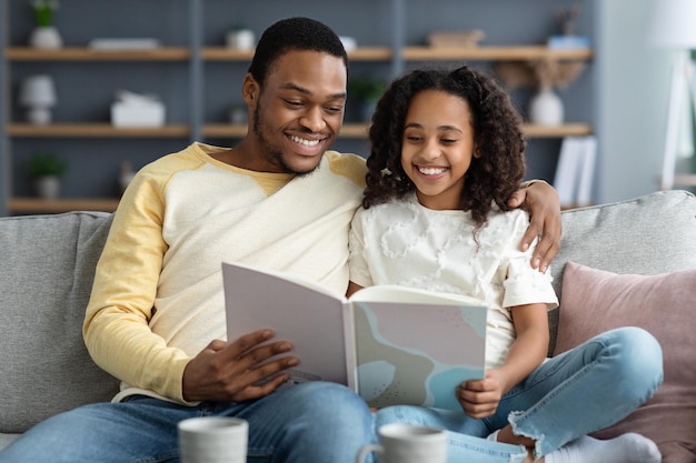 Joyeux père de famille noire et jolie fille lisant un livre ensemble à la maison, père afro-américain et fillette assise sur un canapé dans le salon, profitant d'histoires passionnantes