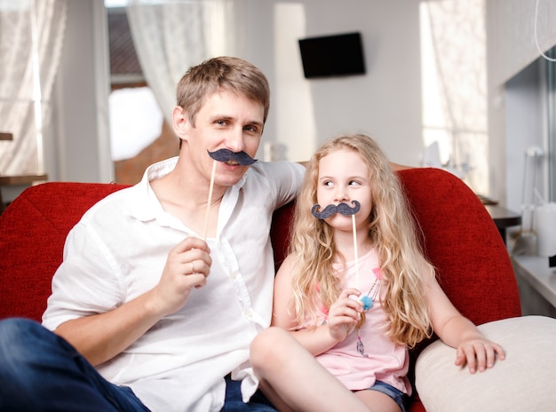 Joyeux père et fille avec moustache artificielle assis ensemble sur une chaise rouge à la maison.