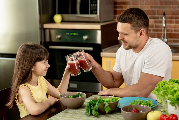 Joyeux père et sa fille prenant le petit déjeuner ensemble dans la cuisine, parler, tenant des verres de jus