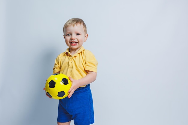 Joyeux petit garçon de 56 ans fan de football dans un t-shirt jaune soutient l'équipe tient un ballon de football dans ses mains isolé sur fond blanc Le concept de loisirs sportifs en famille