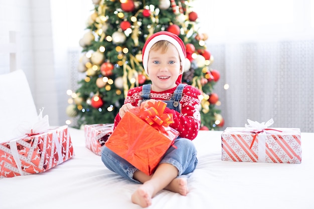 Joyeux petit garçon drôle avec des cadeaux au lit le matin de Noël sur le fond de l'arbre de Noël