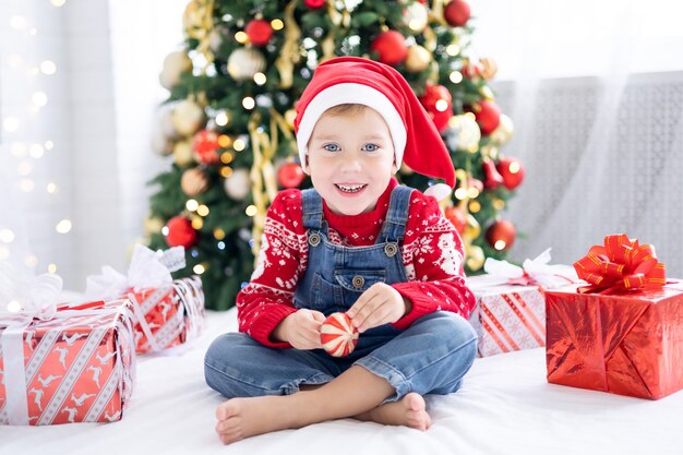 Joyeux petit garçon drôle avec des cadeaux au lit le matin de Noël sur le fond de l'arbre de Noël