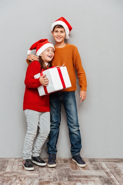 Joyeux petit garçon et fille en chapeaux de père Noël tenant une boîte-cadeau et riant