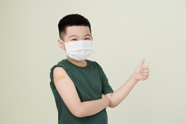 Joyeux petit garçon portant un masque médical et montrant le pouce levé après avoir reçu une injection de vaccin dans le bras