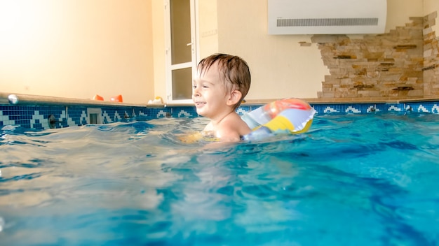 Joyeux petit garçon riant et souriant jouant avec des jouets et apprenant à nager dans une piscine intérieure