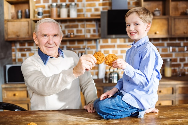 Joyeux petit garçon souriant assis sur la table tout en mangeant des cookies faits maison