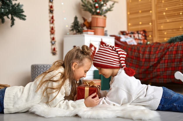 De joyeux petits frère et sœur sont allongés sur le sol dans la chambre de leurs enfants décorée pour Noël et tiennent ensemble une belle boîte-cadeau.