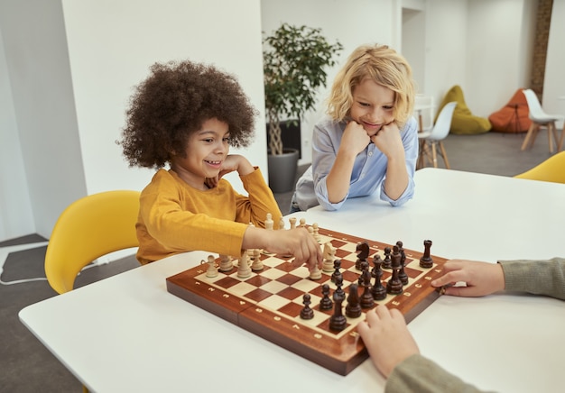 Joyeux petits garçons divers assis à la table et jouant aux échecs à l'école