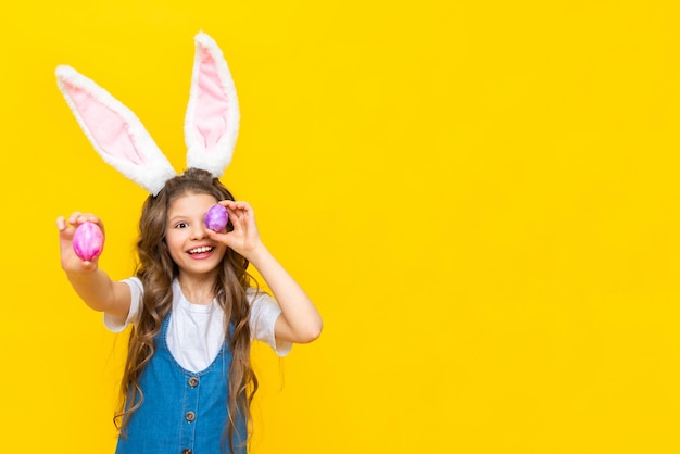 Joyeux printemps Pâques La petite fille ferme les yeux avec des œufs et sourit largement Un charmant enfant avec des oreilles de lapin dans une robe d'été bleue sur un fond jaune isolé Espace de copie