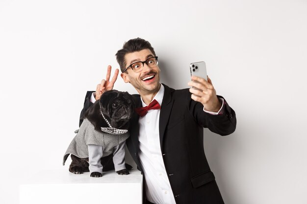 Joyeux propriétaire de chien en costume célébrant Noël avec un chien, prenant un selfie sur un smartphone près d'un mignon carlin noir en costume, fond blanc.