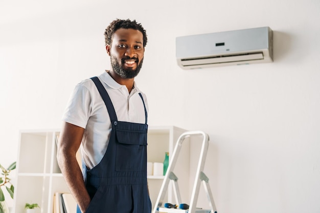 Joyeux réparateur afro-américain debout sur une échelle près du climatiseur et souriant à la caméra