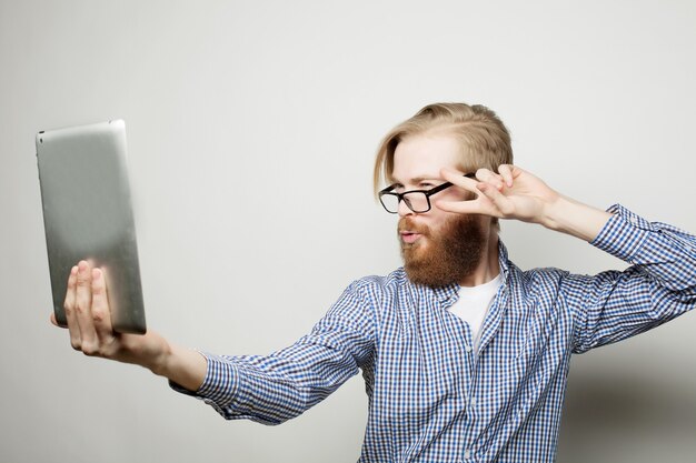 Joyeux selfie. Jeune homme barbu tenant la tablette et faisant la photo de lui-même en se tenant debout sur blanc