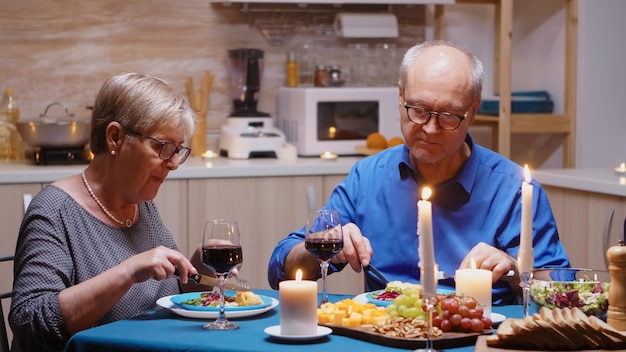 Joyeux vieux couple de retraités amoureux prenant un repas à la maison. Couple d'âge mûr mangeant et discutant pendant un dîner romantique assis à table dans la cuisine moderne, profitant du temps ensemble