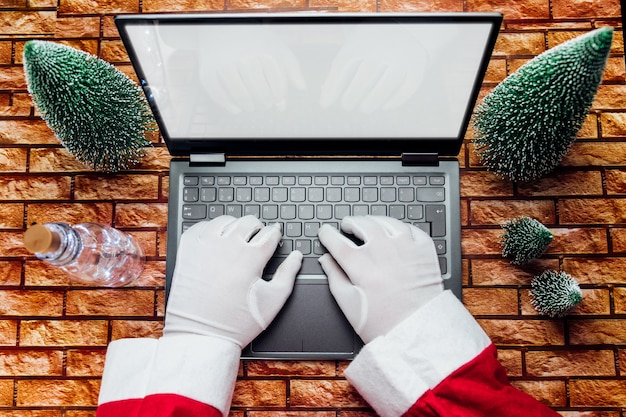 Photo joyeux vieux père noël portant un chapeau tenant une boîte-cadeau à l'aide d'un ordinateur portable assis à la table d'accueil de l'atelier tard le soir du joyeux noël. site web de commerce électronique vacances de temps de noël achats en ligne vente de commerce électronique