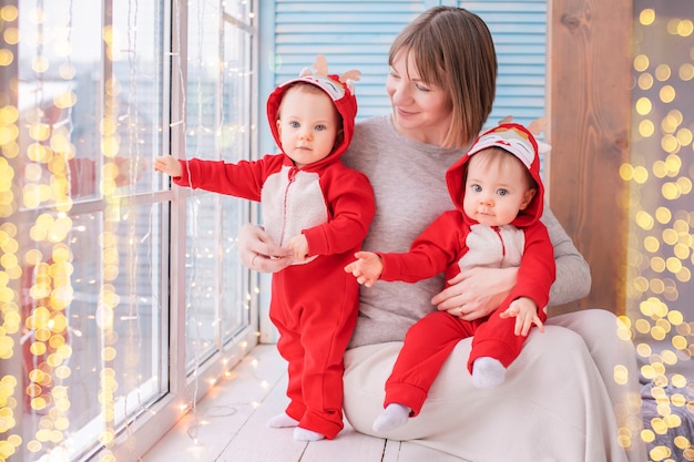 Des jumeaux tout-petits en costumes de père noël de renne rouge sont assis à la maison avec leur mère dans le contexte d'une fenêtre avec des guirlandes