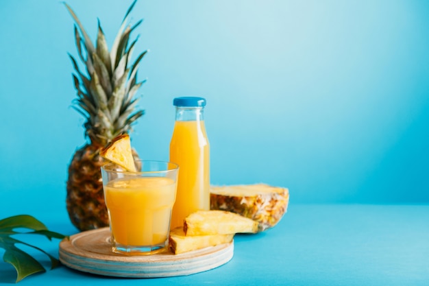 Jus d'ananas savoureux en verre avec des ingrédients, bouteille de jus en verre sur fond d'été de couleur bleue. Cocktail d'ananas naturel frais et jus d'ananas en verre et bouteille avec espace de copie.