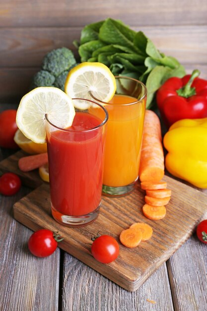 Jus de carotte et de tomate dans des verres et des légumes frais sur une planche à découper sur une table en bois sur fond de mur en bois