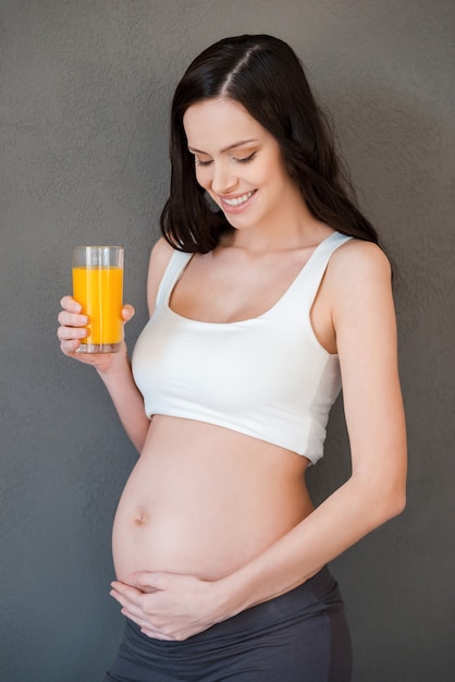 Jus frais pour mon bébé. Belle femme enceinte tenant un verre de jus d'orange et souriant en se tenant debout sur fond gris
