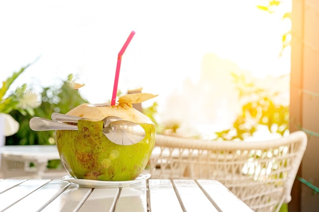 Jus de noix de coco frais avec de la paille et deux cuillères sur une table en bois blanc contre une plage et une montagne floues