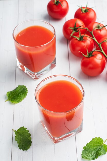 Jus de tomate dans des verres en verre et tomates mûres fraîches sur une branche. Fond en bois blanc avec.