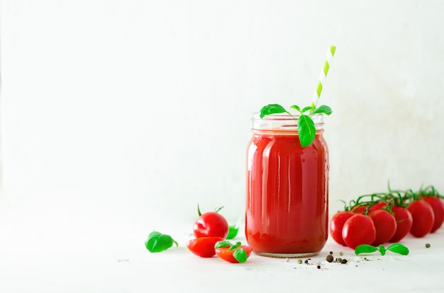 Jus de tomates fraîches biologiques dans un bocal en verre, basilic, cerise, sel, poivre et paille à la lumière.