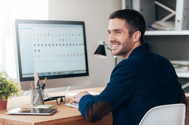 Juste inspiré. Beau jeune homme regardant loin et souriant alors qu'il était assis sur son lieu de travail au bureau